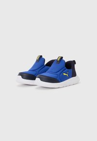 Chaussures slip-on bleues et navy en matériau synthétique, avec une semelle blanche et un logo jaune en accent sur le côté.