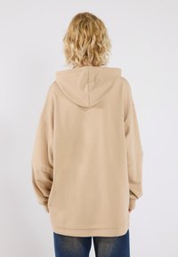 Beige oversized hoodie with a drawstring hood, featuring wide sleeves and a soft fabric texture. Worn with blue jeans.