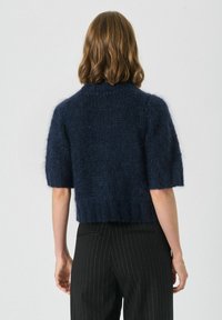 Pulover scurt, de culoare bleu marin, cu mânecă scurtă, realizat dintr-un material tricotat texturat. Are un gât rotund și o croială lejeră, purtat cu pantaloni cu dungi.