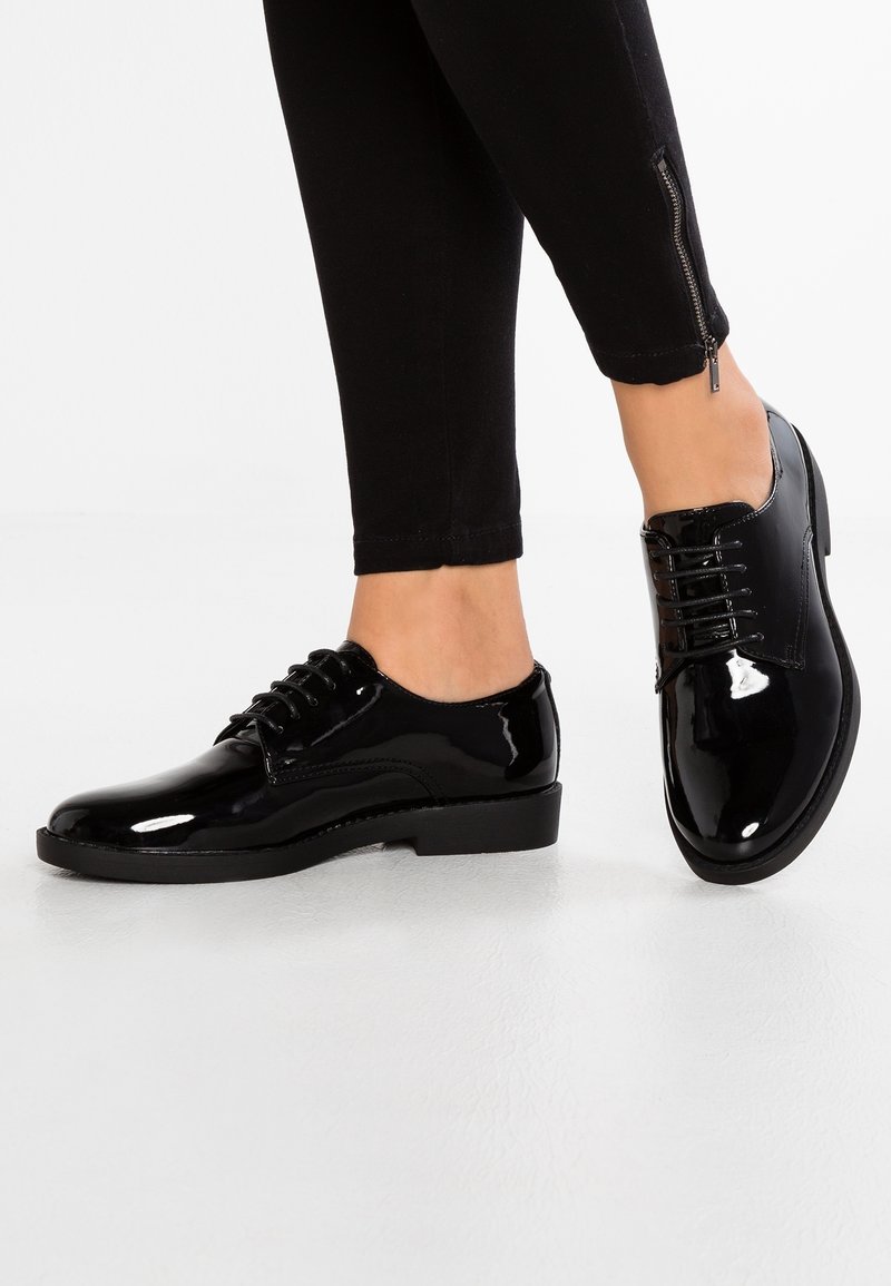 Chaussures à lacets en cuir verni noir avec un bout arrondi, un petit talon et une texture lisse. Présente des lacets noirs et un subtil détail de couture.
