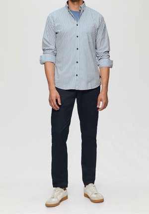 Man wearing a light blue vertical striped button-up shirt, dark pants, light blue undershirt, and white sneakers standing against a plain background.