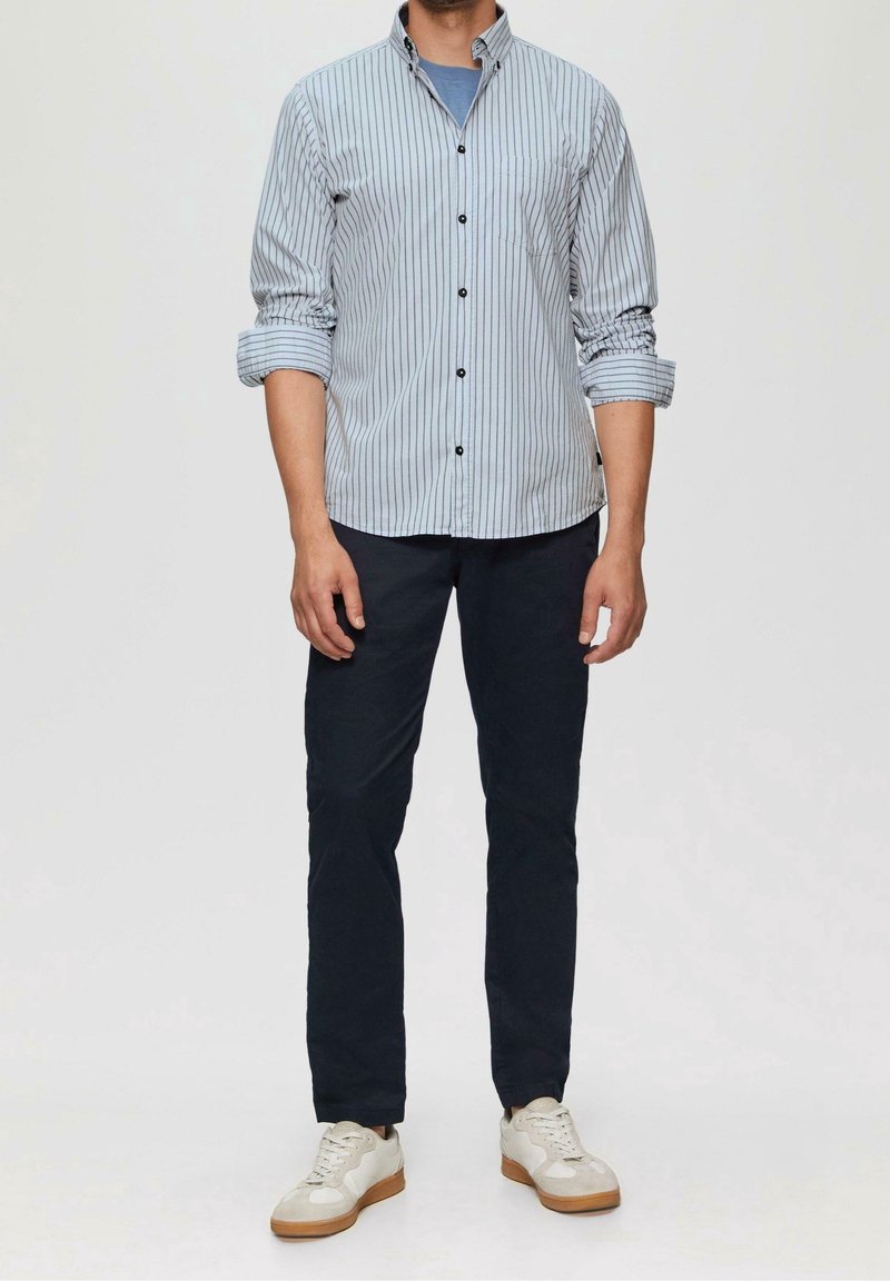 Homme portant une chemise à rayures verticales bleu clair à boutons, pantalon foncé, sous-vêtement bleu clair et baskets blanches, debout devant un fond uni.