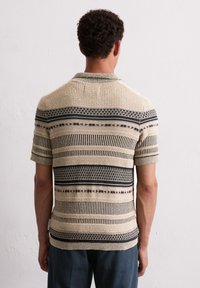 Beżowa koszulka polo z krótkim rękawem, z ciemnoniebieskimi poziomymi paskami i geometrycznymi wzorami; teksturowana tkanina i ściągacz przy kołnierzu.