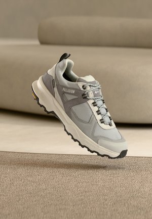 Γκρι και λευκό αθλητικό παπούτσι Palladium με χοντρή σόλα, πάνελ από δίχτυ και σουέτ, να αιωρείται πάνω από μπεζ μοκέτα σε εσωτερικό χώρο.