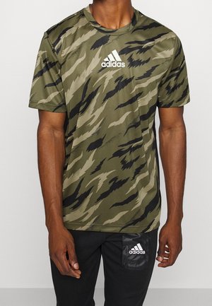 Buste masculin portant un t-shirt Adidas à motif camouflage vert et noir avec un logo blanc, associé à un pantalon noir doté d'une petite étiquette logo Adidas.