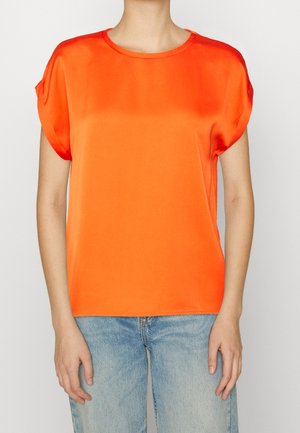 Mujer con blusa de manga corta color naranja brillante y jeans azul claro, de pie contra un fondo claro y liso.