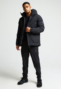 Melnā pūkainā jaka ar kapuci, ar rāvējslēdzēja kabatām un teksturētu audumu. Apvienota ar melnajām sporta biksēm un melniem sporta apaviem.