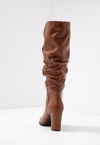 Bota de cuero marrón hasta la rodilla con un diseño arrugado, puntera redonda y un tacón alto grueso. Textura suave con detalles de costura mínimos.