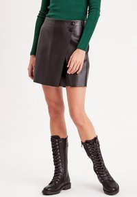 Mini-jupe en cuir noir avec un design enroulé et deux boutons visibles. Le mannequin porte des bottes noires à lacets montantes.