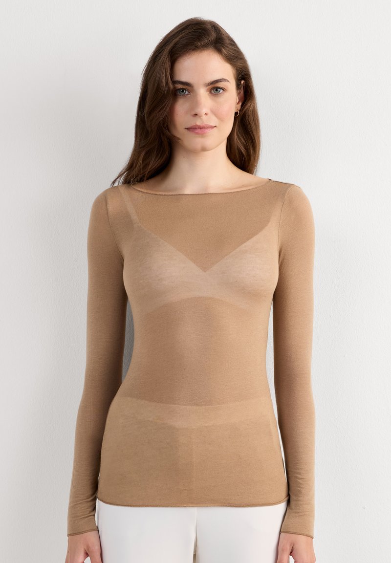 Frau mit langen braunen Haaren, die ein eng anliegendes, durchsichtiges beiges Langarmshirt und weiße Hose trägt, steht vor einem einfarbigen hellen Hintergrund.