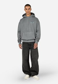 Šedá mikina s predným vreckom a rebrovanými manžetami, s modrou vyšívanou aplikáciou "Sport Club". Skombinovaná s voľnými čiernymi džínsami.