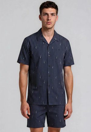 Joven con camisa azul marino de manga corta abotonada y pantalones cortos a juego con rayas verticales blancas y patrones sutiles.