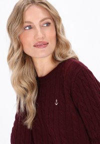 Burgunderfarbener Kabelstrickpullover mit Rundhalsausschnitt und kleinem gesticktem Ankerdetail auf der Brust, das eine weiche Textur zeigt.
