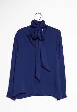 Blouse bleu marine à manches longues avec un col à nœud noué, suspendue sur un cintre noir sur fond blanc.
