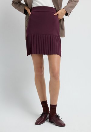 Gonna plissettata bordeaux con vita aderente, che mostra un tessuto testurizzato e un orlo inferiore strutturato. Abbinata a scarpe scure e calze.