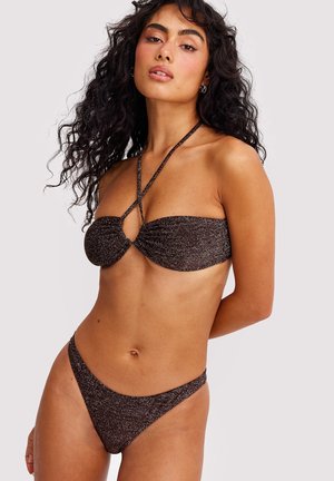 Femme aux longs cheveux bouclés portant un bikini dos-nu pailleté brun foncé, posant avec les bras derrière le dos sur un fond uni.