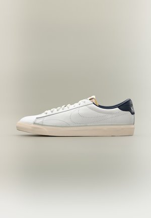 Biela nízka teniska s béžovou podrážkou, námorníckym modrým jazýčkom na päte, bielymi šnúrkami a jemne perforovaným logom Swoosh na boku.