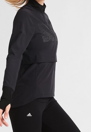 Vrouw die een zwart atletisch shirt met lange mouwen en zwarte Adidas legging met duimgaten draagt, afgebeeld van halverwege de nek tot de bovenbenen tegen een effen achtergrond.