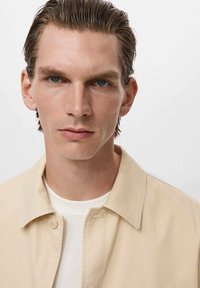 Jachetă bej cu guler peste un tricou alb. Materialul pare neted și croit pe măsură. Modelul are părul scurt, aranjat și ochi albaștri.