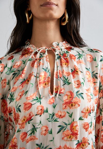 Blusa floral de tejido ligero, con vibrantes flores naranjas sobre un fondo crema, con un escote con lazo y sutiles pliegues en el cuello.