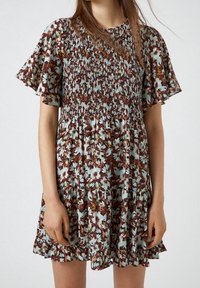 Blommig klänning i brunt, blått och krämfärgat. Har en smockad bodice, fladdrande ärmar och en lager-på-lager volangkant. Lättviktig tyg.