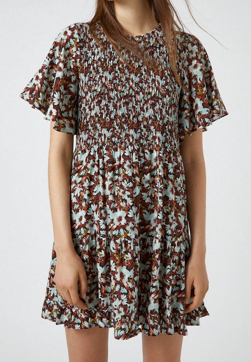 Blommig klänning i brunt, blått och krämfärgat. Har en smockad bodice, fladdrande ärmar och en lager-på-lager volangkant. Lättviktig tyg.