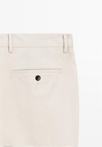 Beige broek met een gladde textuur, voorzien van een knopenluiting aan de voorkant en een enkele achterzak met een zwarte knoopaccent.