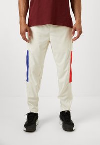 Krem joggebukser med blå og røde sidestykker, tapered fit, glatt tekstur og elastisk midje. Båret med svarte sneakers.