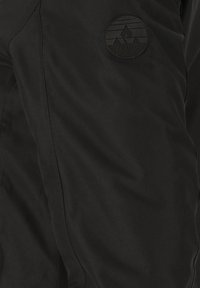 Schwarze Stoffhose mit glatter Textur, ausgestattet mit einem runden, geprägten Logo auf der linken Seite und sichtbaren Nahtdetails.