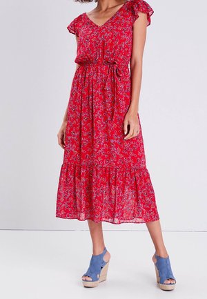 Femme portant une robe midi rouge fleurie avec manches courtes et ceinture à la taille, associée à des sandales compensées bleues à bout ouvert.