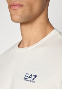 Balta medvilninė marškinėliai su apvalia iškirpte, su mėlynu EA7 Emporio Armani logotipu ant krūtinės, lygaus paviršiaus ir prigludusio kirpimo.
