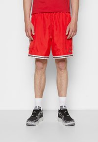 Czerwone spodenki sportowe z białymi paskami po bokach i małym czarnym logo. Noszone z czarnymi tenisówkami o fakturowanym designie i białymi skarpetkami.