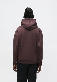 Felpa con cappuccio color marrone scuro con cappuccio ampio e regolabile, spalle larghe e taglio dritto. Tessuto morbido in misto cotone con polsini a coste.