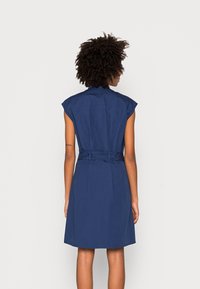 Marineblaues Kleid mit kurzen Kappärmeln, tailliert mit einem Gürtel. Verfügt über einen klassischen Kragen und reicht bis knapp über das Knie. Glatter Stoff.