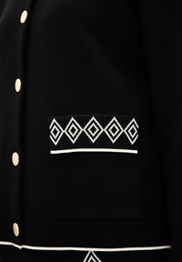 Svart cardigan med gyllene knappar, en texturerad yta och en vit diamantmönstrad ficka samt ett accentband nära fållen.