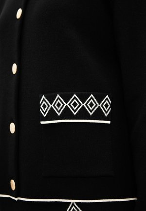 Schwarze Strickjacke mit goldenen Knöpfen, einer strukturierten Oberfläche und einer weißen Rauten-muster-Tasche sowie einem Akzentband am Saum.