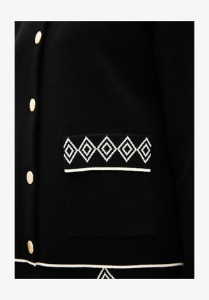 Schwarze Strickjacke mit goldenen Knöpfen, einer strukturierten Oberfläche und einer weißen Rauten-muster-Tasche sowie einem Akzentband am Saum.