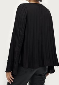 Une femme portant un haut noir plissé à manches longues avec des manches évasées et un pantalon en cuir noir, vue de dos.