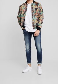 Hombre vistiendo una chaqueta bomber de estampado colorido sobre una camiseta blanca, con jeans azules remangados y zapatillas blancas, de pie contra un fondo sencillo.