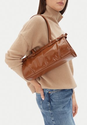 Donna che indossa un maglione a collo alto beige e jeans blu, con una borsa in pelle marrone lucida appesa alla spalla su sfondo bianco.