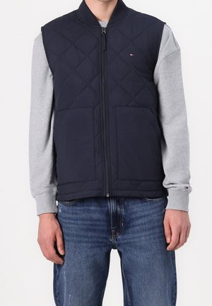 Mann trägt eine marineblaue gesteppte Weste mit Reißverschluss über einem hellgrauen Langarmshirt und blauen Jeans, steht vor einem schlichten weißen Hintergrund.