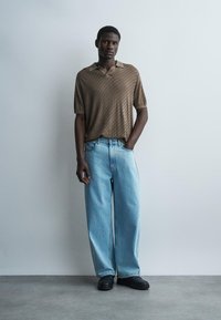 Mann iført brun teksturert kortermet poloshirt, lyseblå vide jeans og svarte sko, stående foran en ensfarget lys bakgrunn.