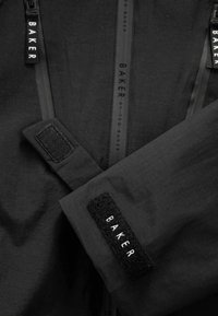 Schwarze Jacke mit glatter Textur, ausgestattet mit kontrastierenden schwarzen Reißverschlüssen und einem Klettverschluss-Patch mit der Aufschrift "BAKER" am Ärmel.