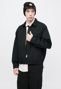 Μαύρο μπουφάν με φερμουάρ και γιακά, με ελαστική μέση, φορεμένο πάνω από ένα ανοιχτό ροζ πουκάμισο και μαύρη γραβάτα· συνδυασμένο με μαύρο σκούφο.