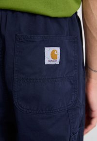 Pantaloni scurți cargo bleumarin, fabricati dintr-un material rezistent, cu o mare buzunar pe picior și un eticheta albă cu un logo portocalie pe spate.