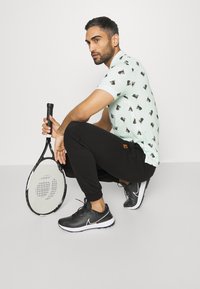 Világos zöld póló fekete kutyás mintával, fekete sportnadrág és fekete Nike tornacipő. Teniszütőt tart a kezében, ülő pozícióban.