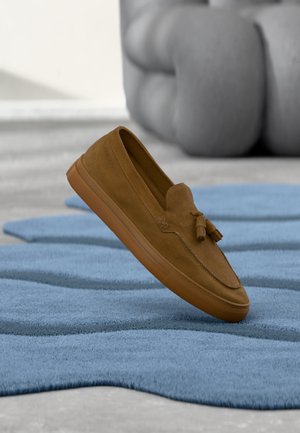Brown suede loafer with tassels floating above textured blue rug and blurred gray sculpture in background.