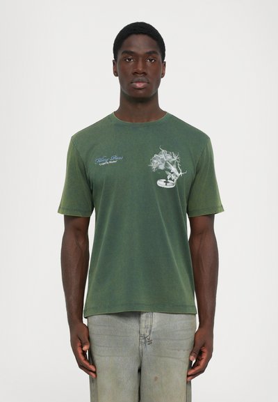 Mand iført en falmet grøn t-shirt med hvid blomster- og tekstdesign på brystet, parret med lysegrå jeans, stående imod en ensfarvet baggrund.
