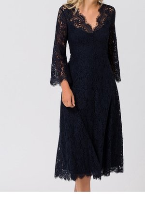Femme portant une robe en dentelle noire longueur genou avec des manches trois-quarts et un décolleté en V festonné, debout sur un fond uni.