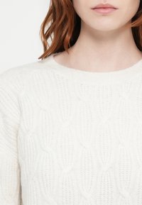 White cable-knit sweater with a round neckline, featuring a textured pattern of intertwining loops and ribbed cuffs. Soft, lightweight material.
